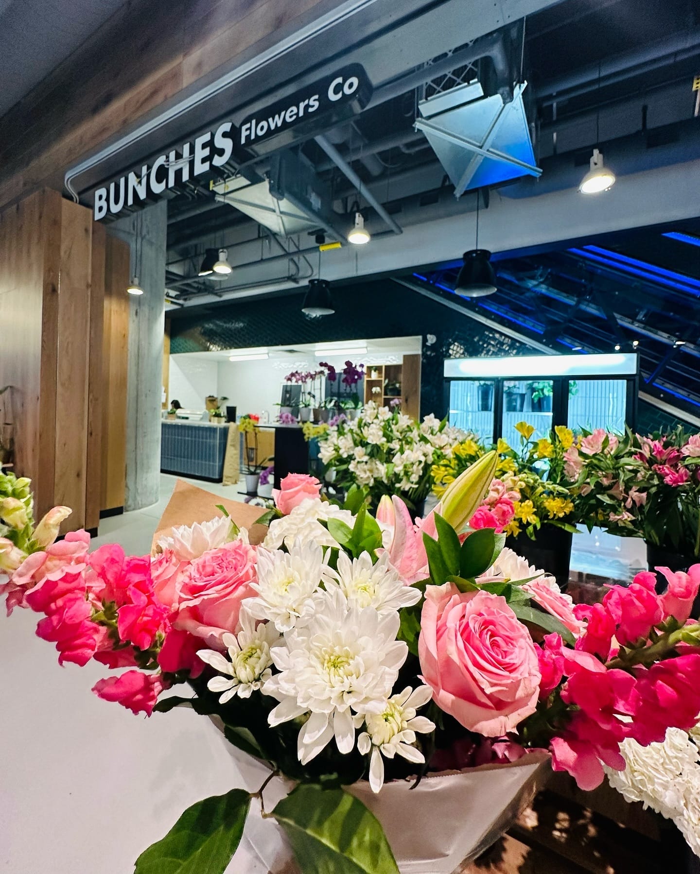 A colorful bouquet of pink and white flowers inside Bunches Flowers Co. at The Well, with the store's sign in the background.