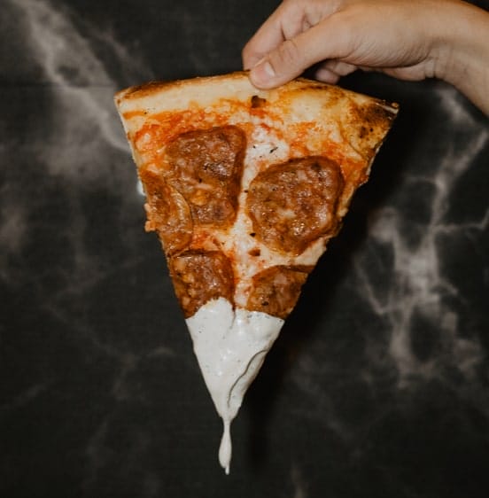 The image depicts a single slice of pizza being held up, with a rich cheese and pepperoni topping. The crust looks golden and crispy, while the cheese stretches as it melts from the slice. The sauce has a slightly rustic, tomato-heavy look, paired perfectly with the generous pepperoni slices. The drip of sauce and cheese from the bottom of the slice adds to its appeal, suggesting this could be from a pizzeria known for its indulgent, hearty slices. The overall vibe is a classic, comforting pizza experience.