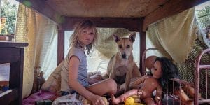 A young girl sits inside a makeshift fort under a table, surrounded by dolls and toys, alongside a loyal dog. The scene is bathed in soft natural light, capturing a sense of innocence and imagination amidst a backdrop of makeshift living conditions. The girl's gaze is thoughtful and serious, hinting at the deeper emotional journey she is on in Don't Let's Go to the Dogs Tonight.