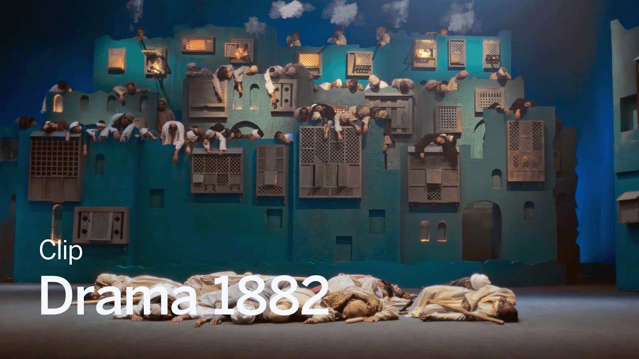 A still from Drama 1882 showcasing a surreal, theatrical scene with a large blue structure resembling a cityscape. People are draped across windows and balconies, appearing limp, while others lie on the ground below, creating a striking, dreamlike image. The set design features intricate, lattice-like windows and soft lighting, evoking a sense of historical and artistic depth. The composition suggests a haunting and otherworldly atmosphere, capturing the visual richness of the film.