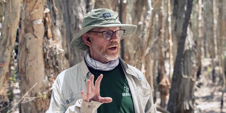 A photo of director Ron Howard standing in a forest setting, wearing a wide-brimmed hat, glasses, and outdoor gear. He appears to be giving directions or discussing something important, with a calm yet focused expression. The background is filled with tall trees, enhancing the natural environment, suggesting this image was taken during the filming of an outdoor project.