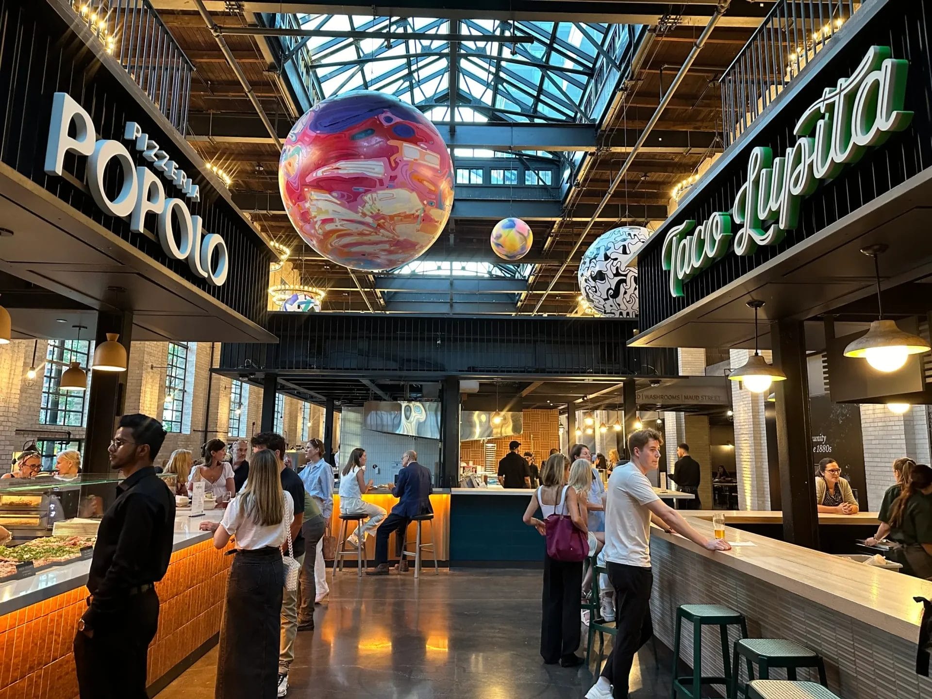 Interior of Waterworks Food Hall in Toronto's King West district, featuring bustling food vendors such as Pizzeria Popolo and Taco Lupita. The vibrant space showcases a modern, industrial design with hanging globe-like art, large windows, and a lively dining atmosphere.