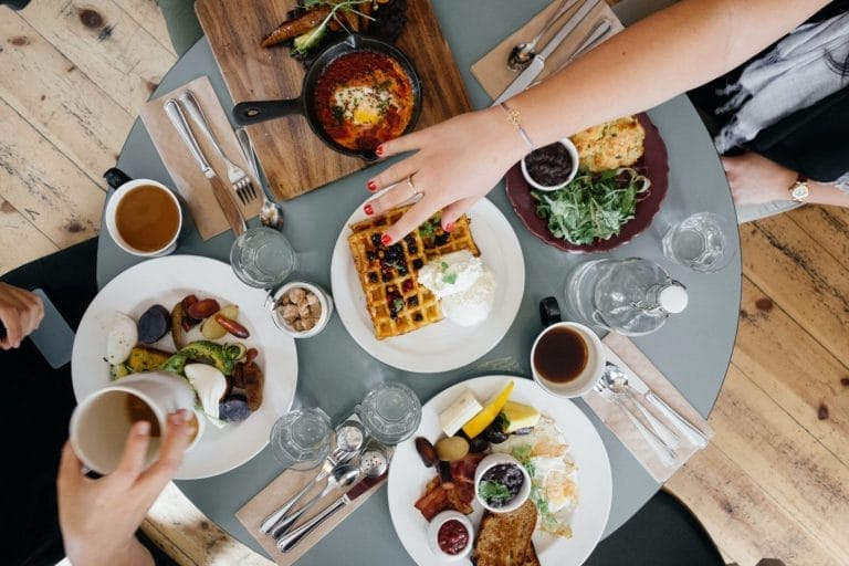 This image captures a cozy brunch scene, with a variety of dishes laid out on a rustic wooden table. The spread includes fluffy waffles topped with whipped cream and berries, a vibrant shakshuka in a cast iron skillet, fresh greens, and a plate of breakfast sides like roasted potatoes, sausage, and perfectly cooked eggs. Cups of coffee accompany the meal, creating an inviting, social atmosphere. The overall setting suggests a casual yet sophisticated brunch, ideal for a relaxed gathering with friends.