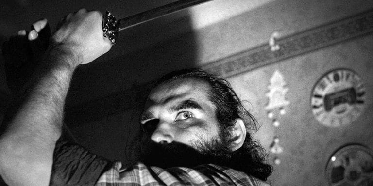 A man grips a sword with intense determination, his eyes wide and focused, suggesting a moment of tension or preparation for an action. The black-and-white image enhances the sense of drama and raw emotion, highlighting his expression in sharp contrast to the surrounding environment.