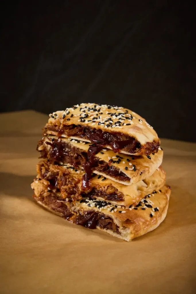 Close-up of a stacked Naan filled with tender pulled meat, drizzled with BBQ sauce, and topped with black and white sesame seeds at Waterworks Food Hall in Toronto. This savory treat is served by Karak, known for its fusion of traditional South Asian flavors with modern twists.