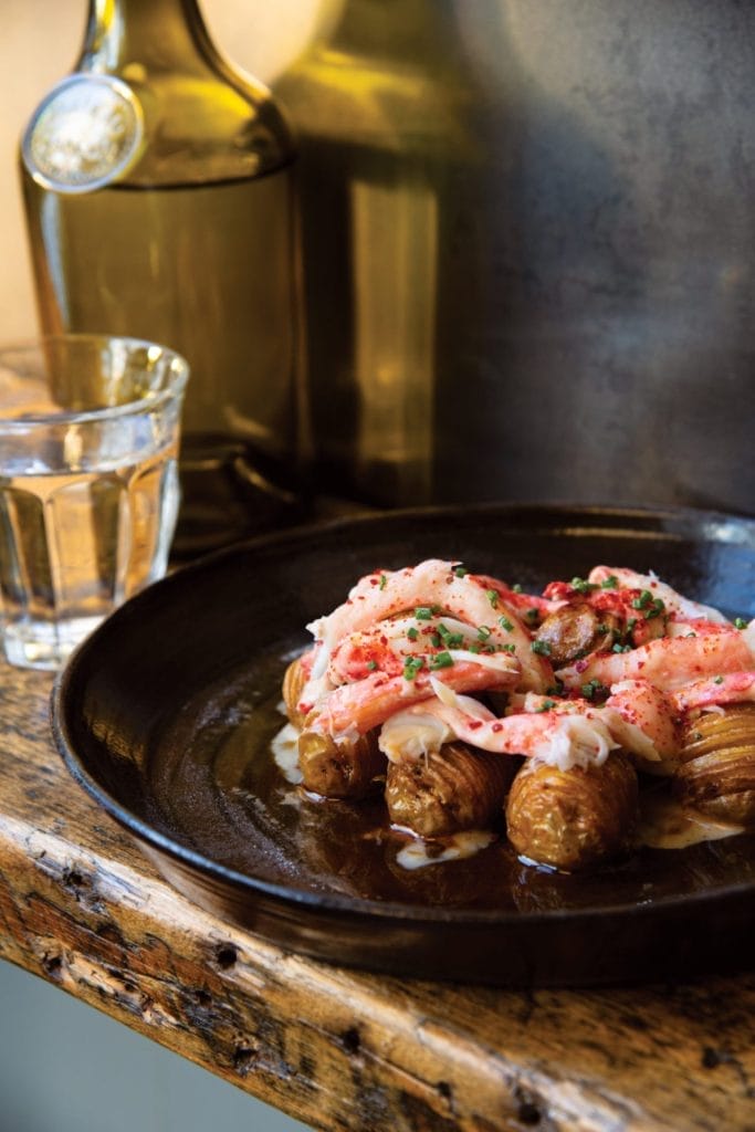 A close-up of a gourmet dish featuring crispy roasted potatoes topped with succulent crab meat, garnished with finely chopped herbs, served on a rustic dark ceramic plate. A glass of clear drink and a bottle are blurred in the background, placed on a wooden table with a rustic texture.