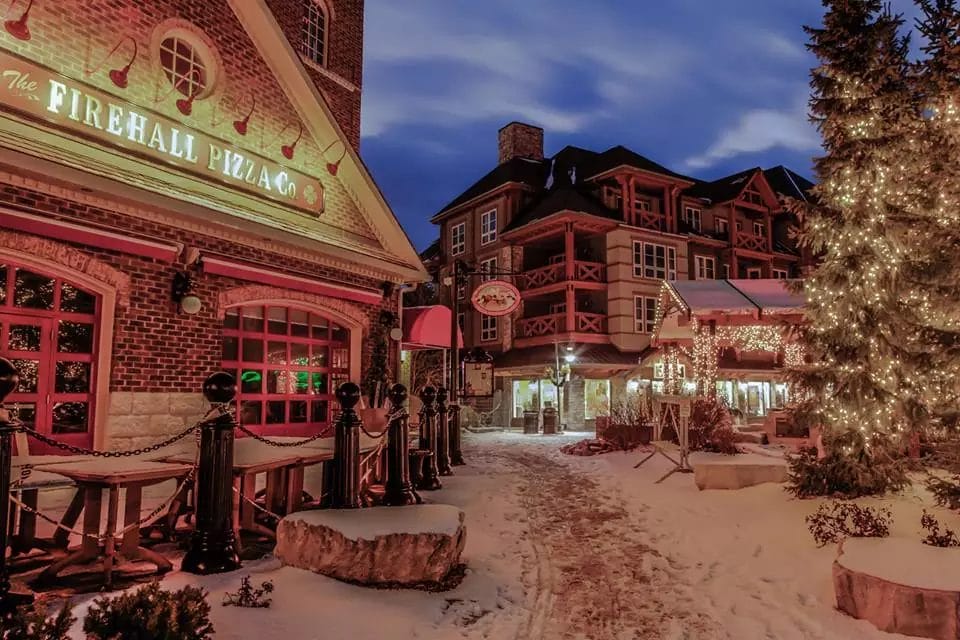 A cozy winter evening at Blue Mountain Village, featuring The Firehall Pizza Co. with snow-covered pathways and warmly lit buildings. A large Christmas tree adorned with festive lights adds to the holiday charm of the scene. The rustic architecture and inviting atmosphere make it a perfect winter retreat for visitors enjoying the holiday season.