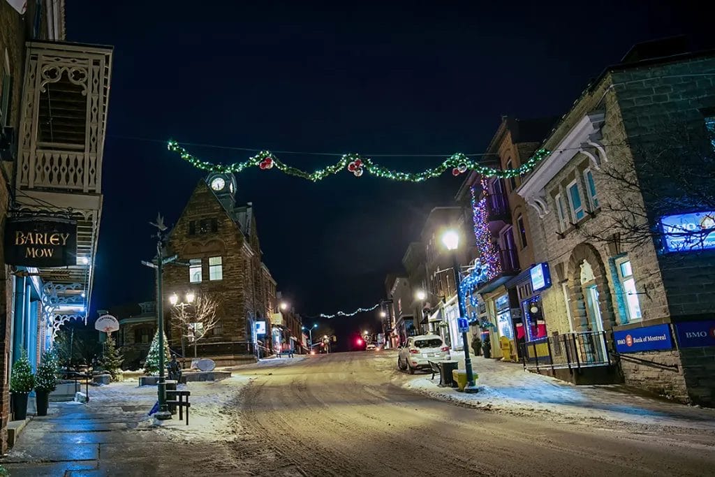 A peaceful small-town street decorated for Christmas with garlands and lights strung overhead, snow-covered sidewalks, and charming, illuminated storefronts. The street appears quiet, evoking a cozy holiday atmosphere.