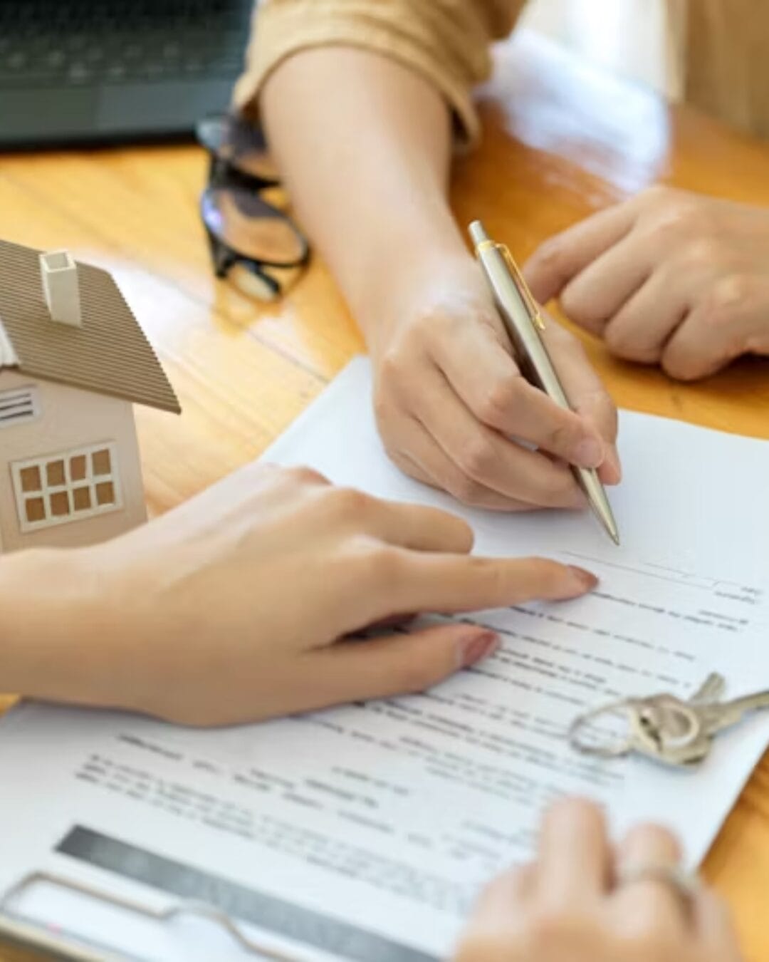 Two people review and sign a real estate contract on a wooden table. A small model house, a pair of keys, and a clipboard with documents are visible.