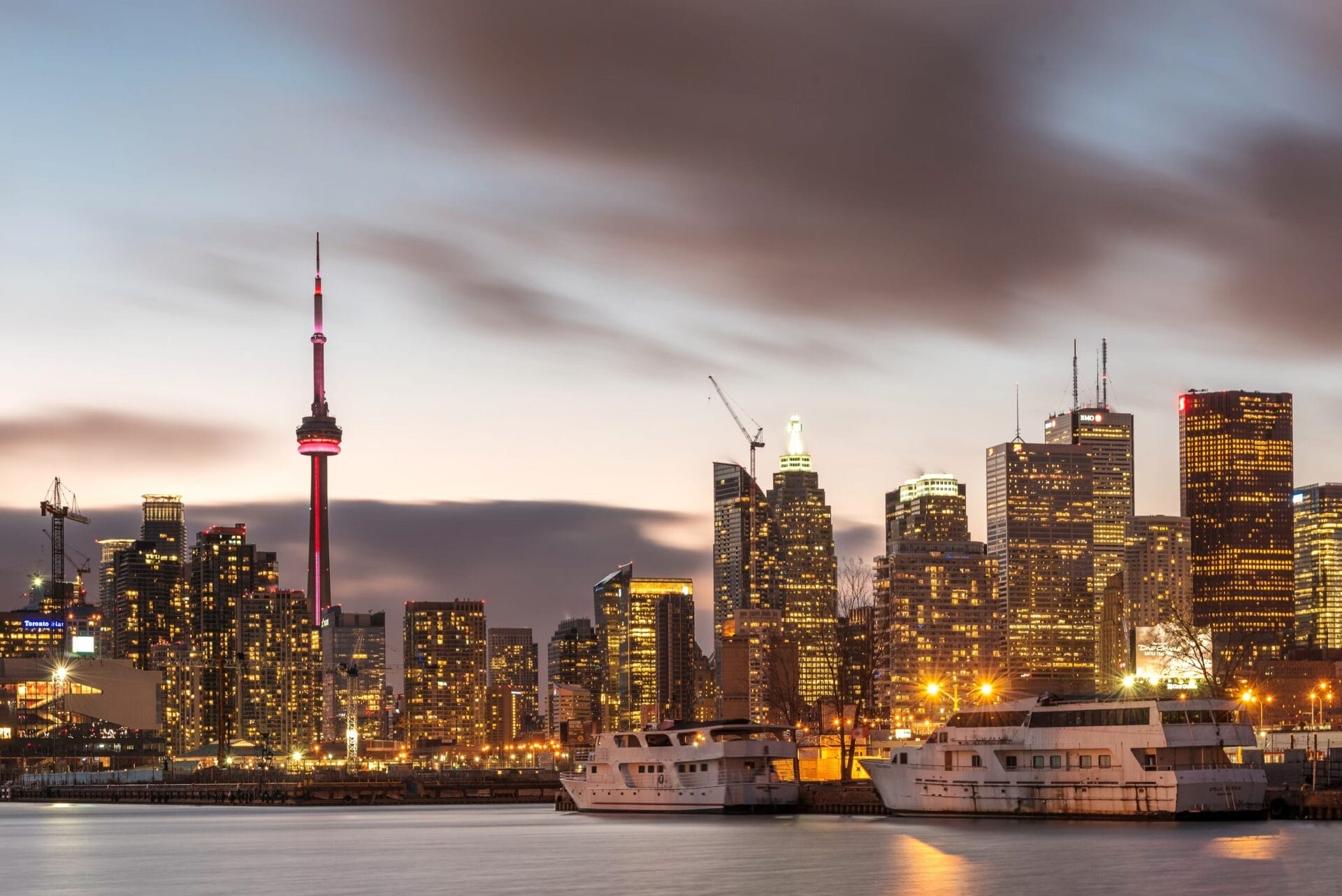 Toronto skyline at dusk with the CN Tower illuminated in red, high-rise buildings glowing with lights, and boats docked along the waterfront.