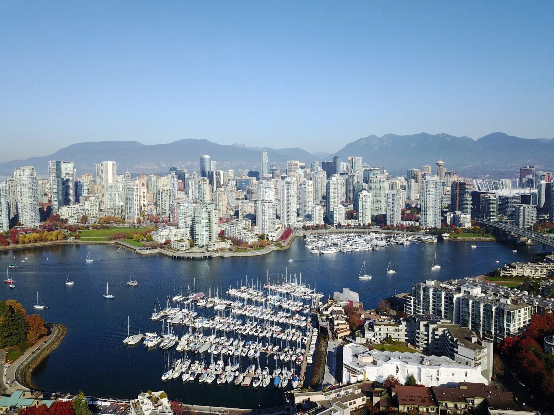 Aerial view of Vancouver with high-rise buildings, a marina filled with boats, and mountains in the background under a clear blue sky.