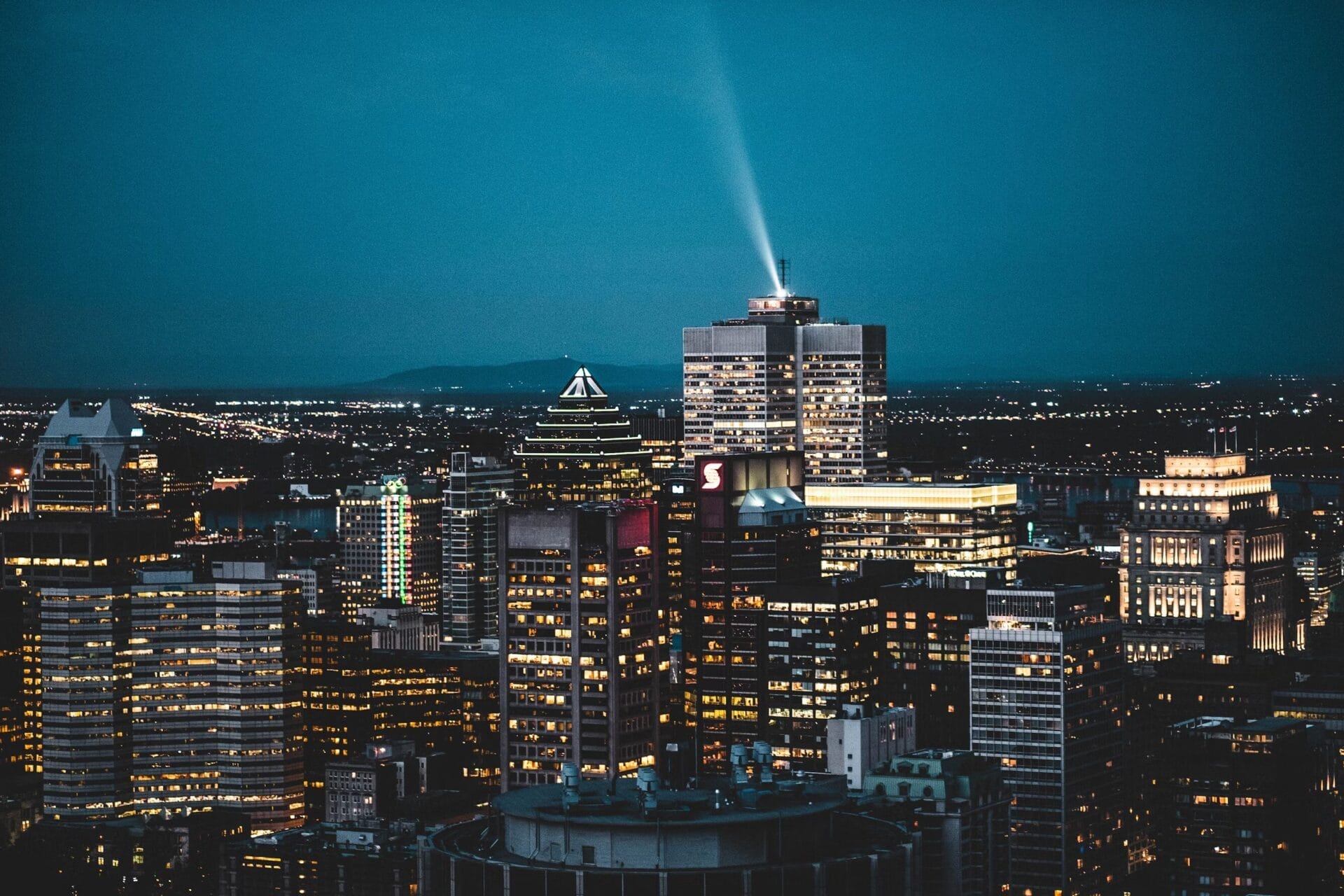 Nighttime view of downtown Montreal, Quebec, featuring illuminated skyscrapers and a spotlight beaming from the top of a high-rise building against a dark blue sky.