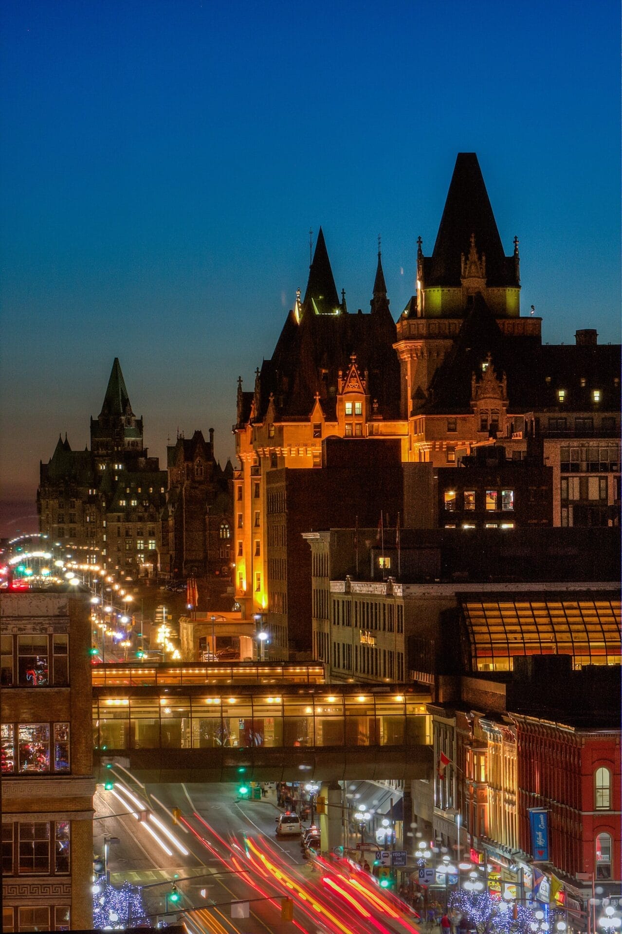Evening view of downtown Ottawa, Ontario, featuring the illuminated Fairmont Château Laurier with its turrets and warm lighting. The foreground shows a busy street with streaks of light from moving vehicles and festive decorations.