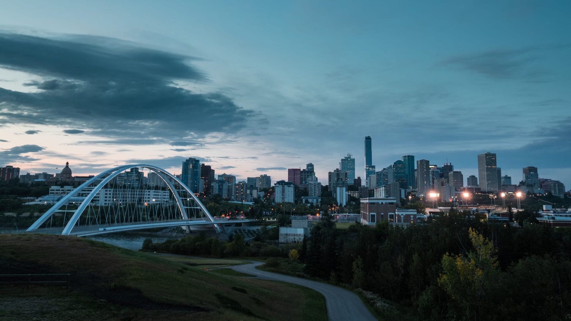 Dusk view of Edmonton, Alberta, featuring the Walterdale Bridge with its white arches spanning the North Saskatchewan River. The city skyline rises in the background under a cloudy sky, with streetlights and buildings beginning to glow.