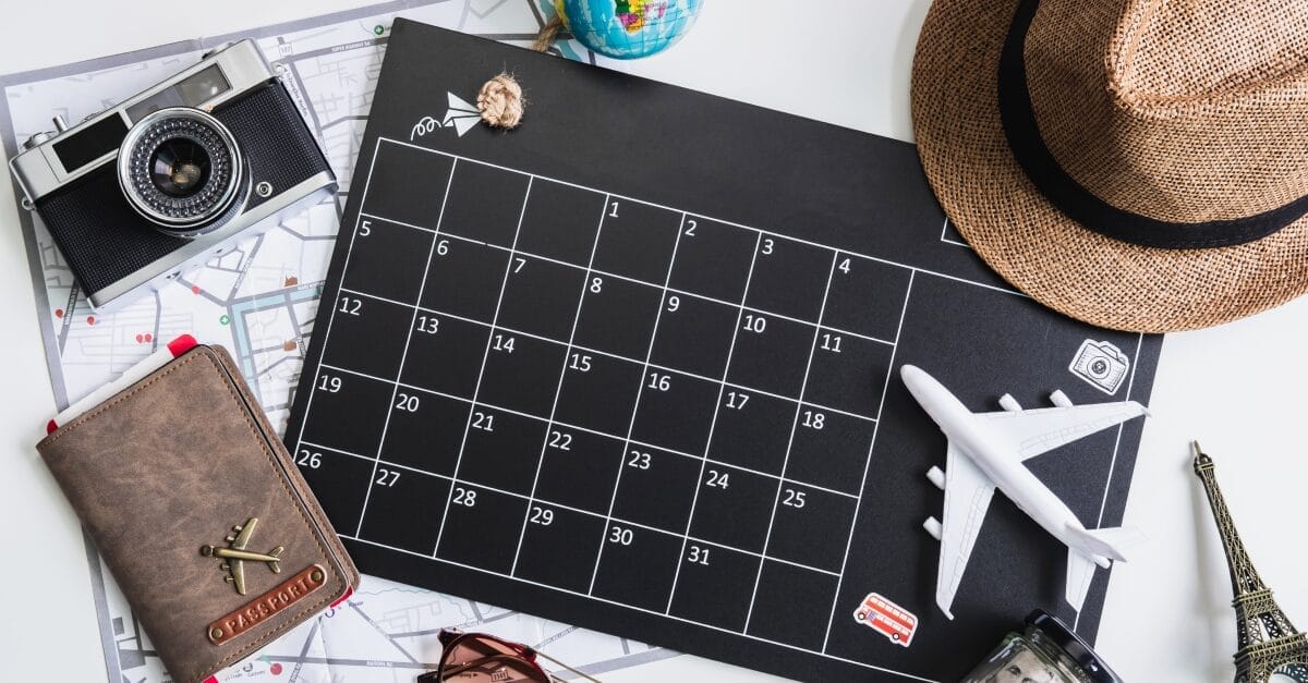 Flat lay photograph featuring travel essentials arranged around a black calendar. Items include a vintage camera, a world map, a passport with an airplane emblem, a straw hat, a miniature airplane model, sunglasses, and a small Eiffel Tower souvenir, symbolizing travel planning and adventure.