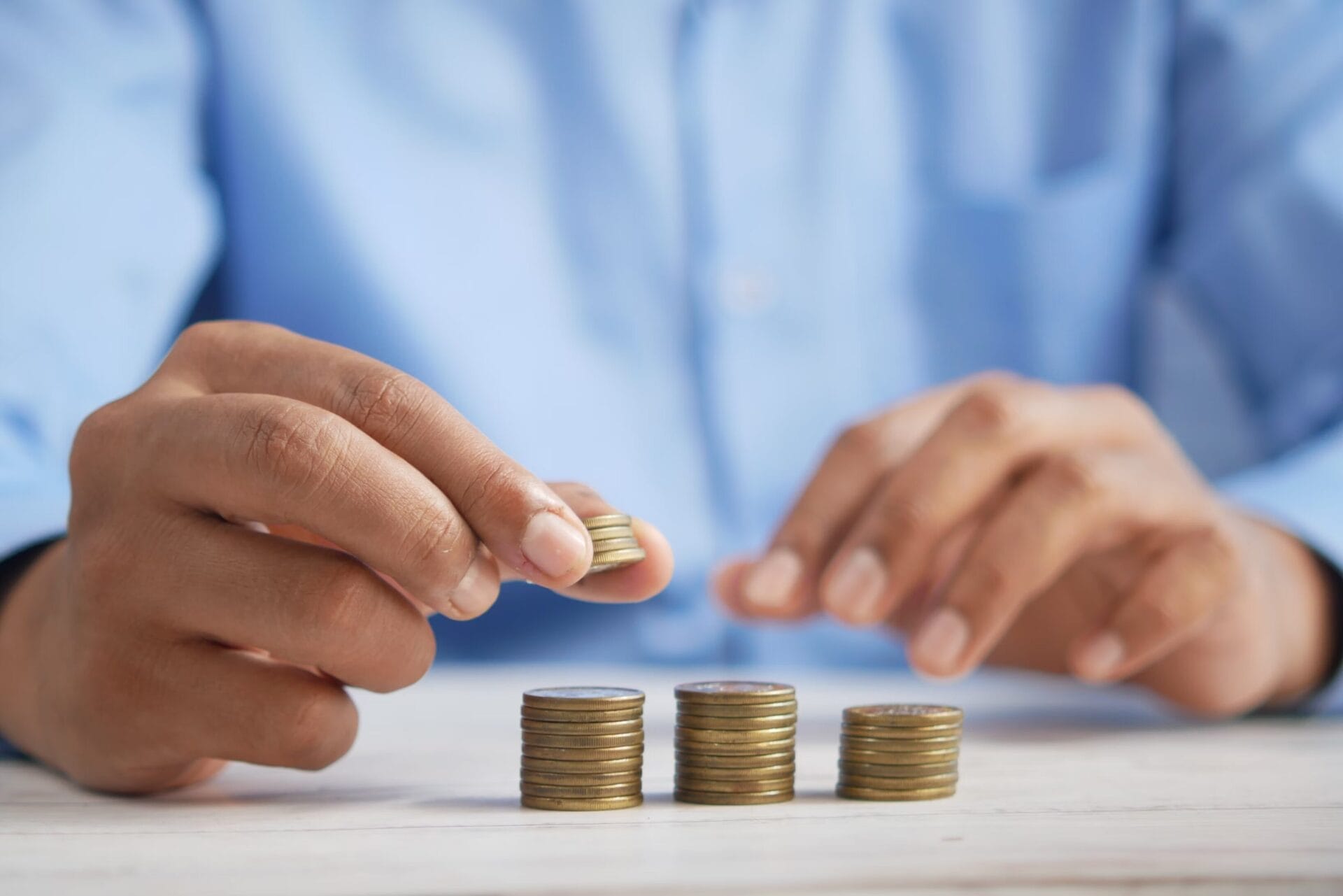 A person in a blue shirt stacking coins into neat piles on a wooden surface, representing savings or financial planning.