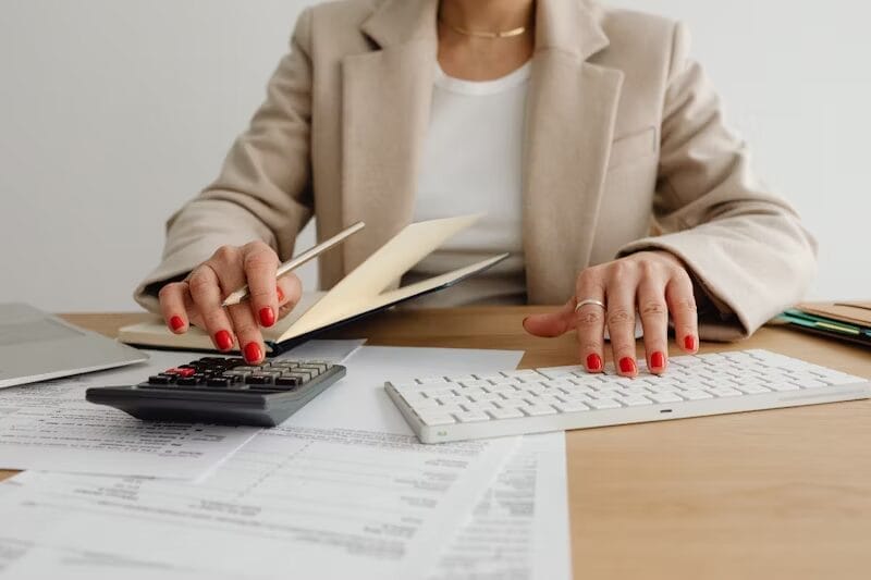 A woman wearing a beige blazer and red nail polish works at a desk with financial documents, a calculator, a keyboard, and a laptop. She holds a pencil while using the calculator, appearing focused on calculations.