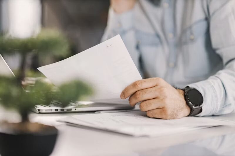A man wearing a light denim shirt and a black wristwatch reviews a document at a desk with papers, a laptop, and a small potted plant. The focus is on his hands and the paper, while the background is blurred.