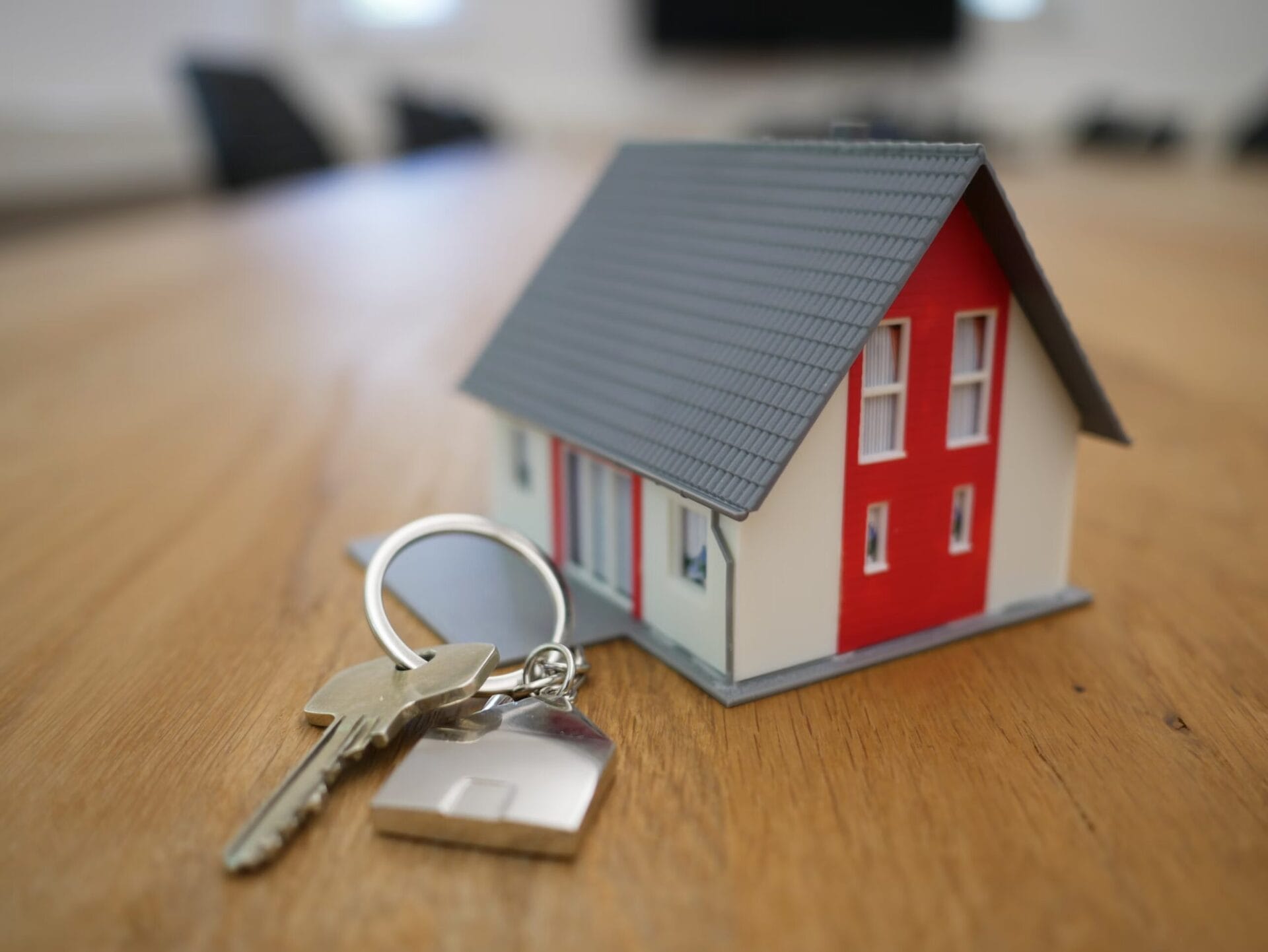 A set of house keys with a house-shaped keychain lies on a wooden table next to a small model of a two-story house with a red and white exterior and a gray roof. The background is blurred, suggesting an indoor office or meeting space.
