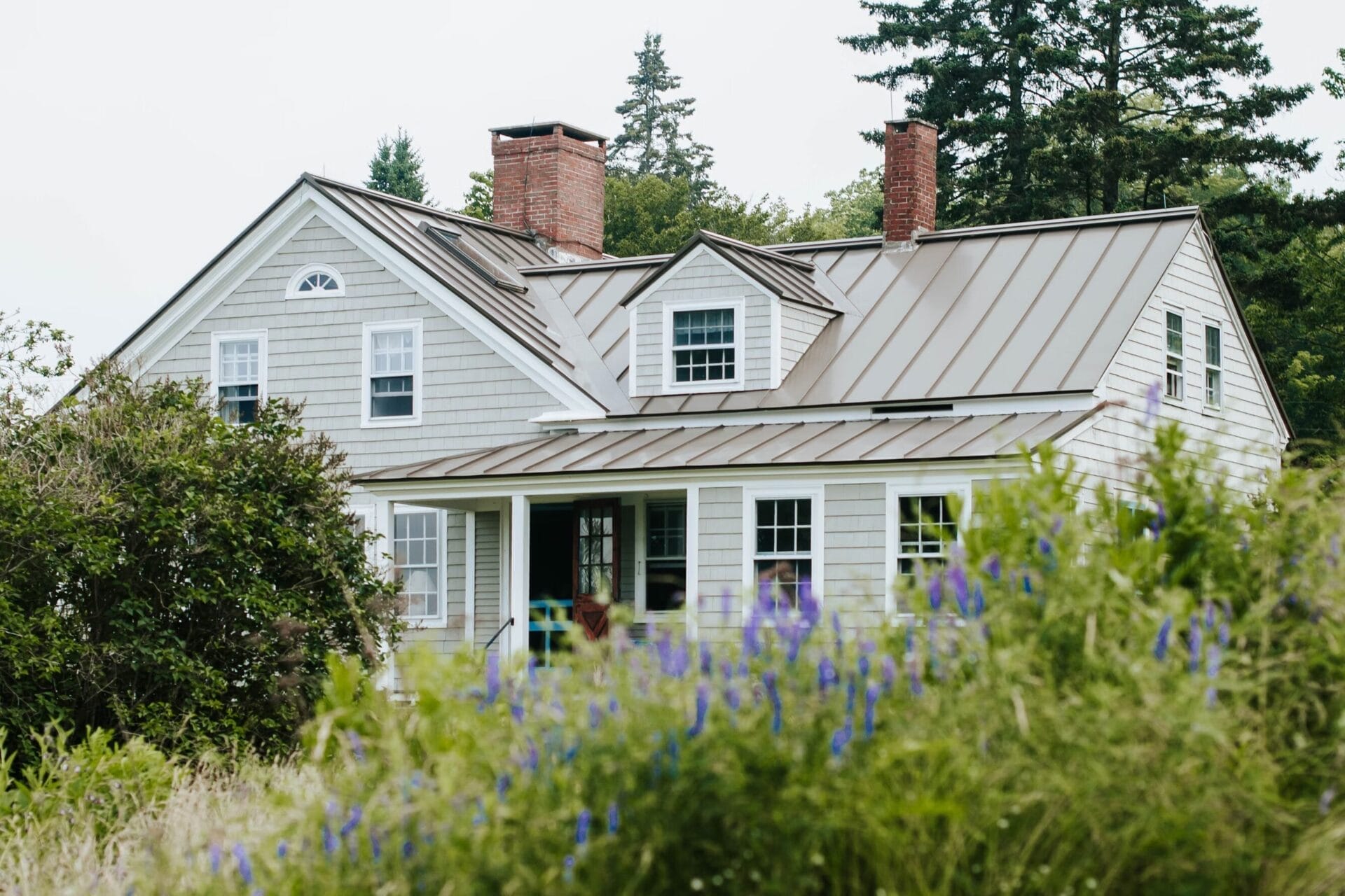 A grey country-style house with a metal roof and brick chimneys sits among lush greenery and purple wildflowers. Tall trees stand in the background, framing the home. | Buy a home