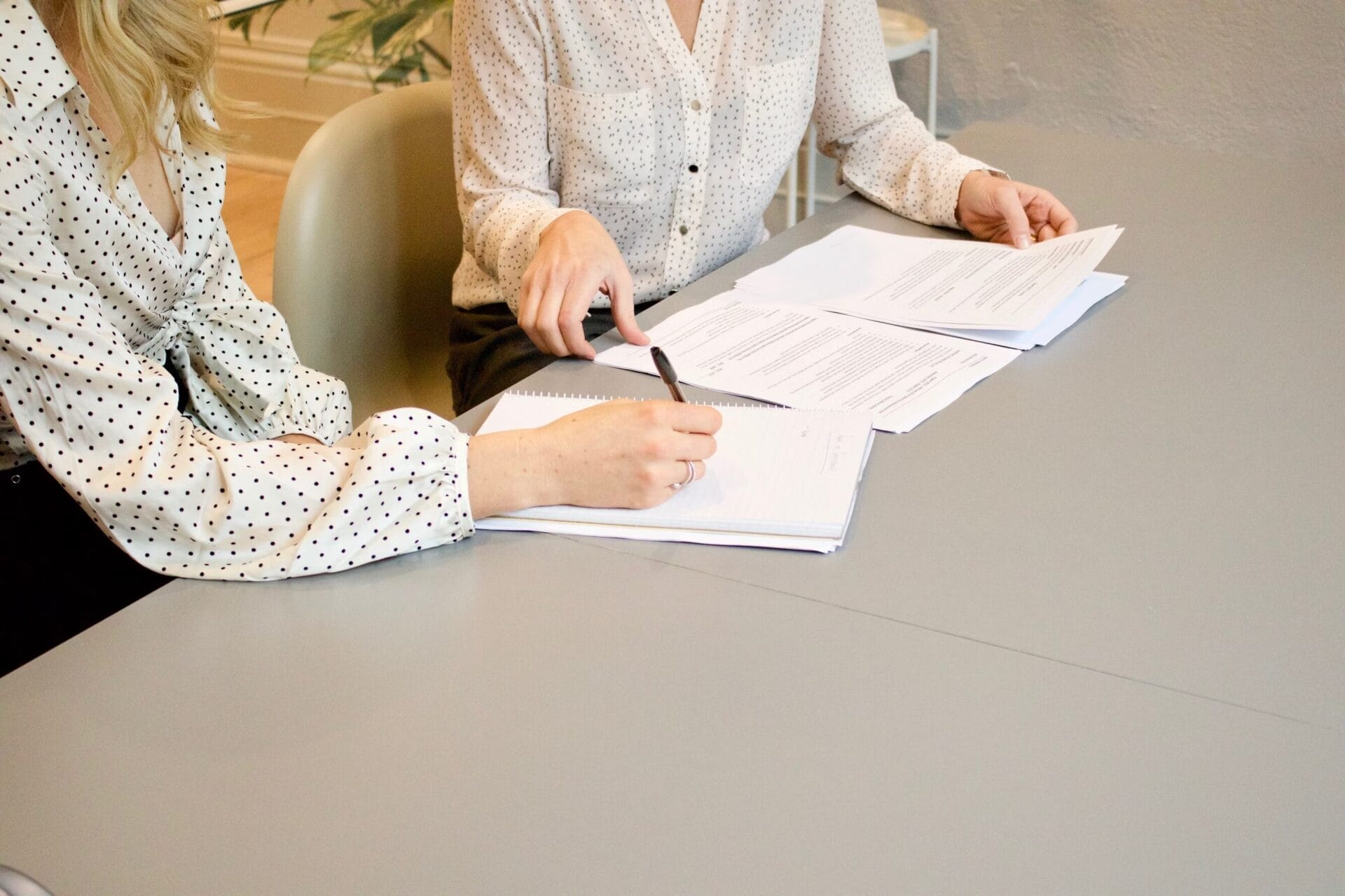 Two people in polka-dotted blouses are seated at a table, reviewing documents together. One is holding a pen and taking notes while the other points at the papers.