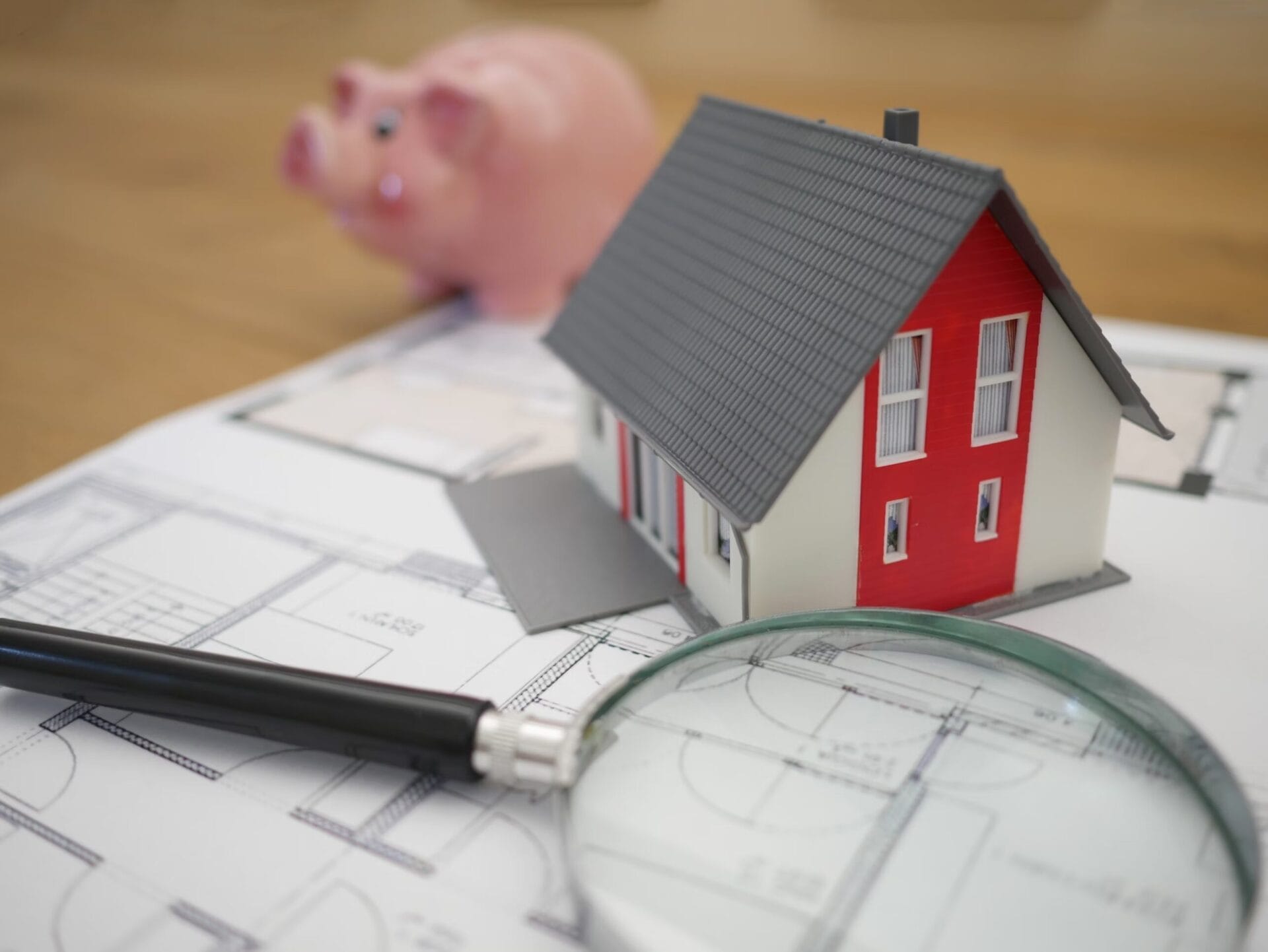 A small model house sits on top of architectural blueprints, accompanied by a magnifying glass and a pen. A pink piggy bank is visible in the background.