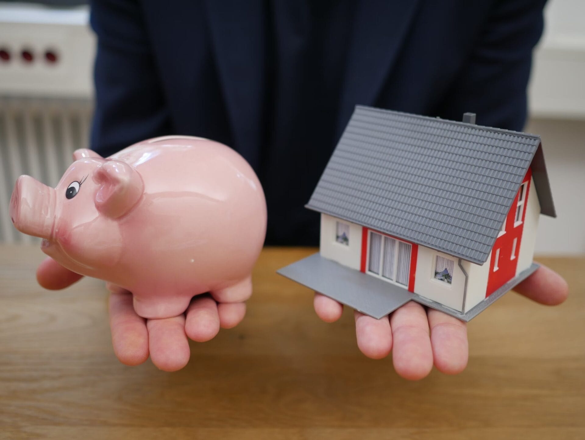 A person in a dark suit holds a pink piggy bank in one hand and a small model house in the other. The image represents financial decisions related to saving and homeownership.