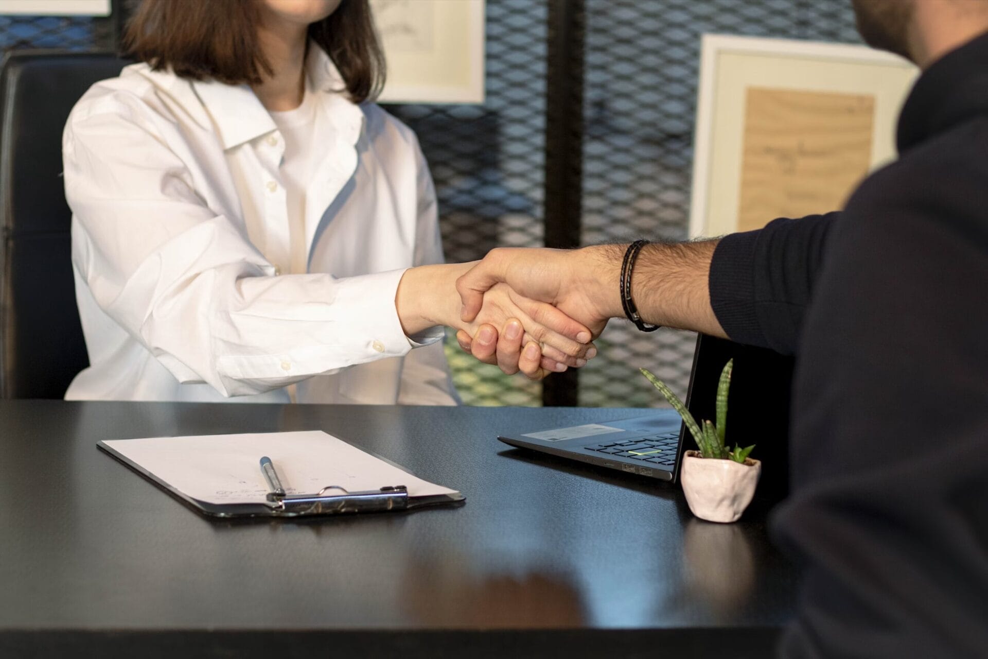 A woman in a white shirt and a man in a dark sweater shake hands across a desk, suggesting a business agreement. A clipboard, pen, laptop, and small potted plant sit on the desk. | Buy a home