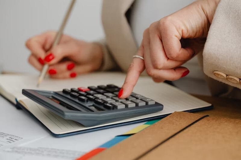 A close-up of a person using a calculator while taking notes in a notebook. The individual is wearing a beige blazer and has red nail polish, with documents and folders visible on the desk.