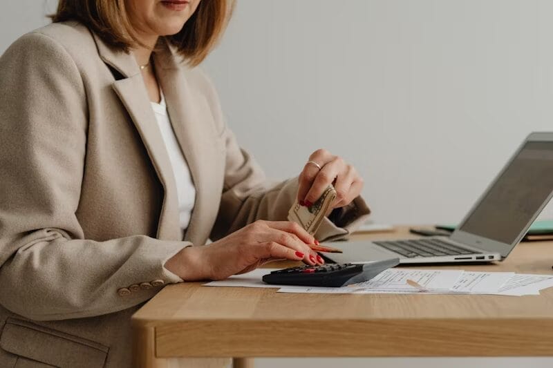 A woman in a beige blazer holds cash in one hand while using a calculator with the other. She is seated at a desk with financial documents, a laptop, and a pen.