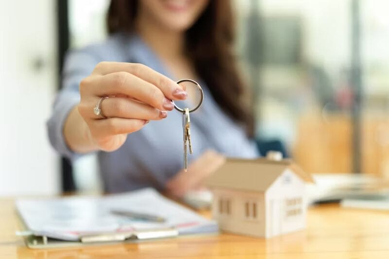 A woman in a blue blouse extends a keyring with house keys, symbolizing a real estate transaction. A small model house, clipboard, and pen are on the table in front of her. | Buy a home