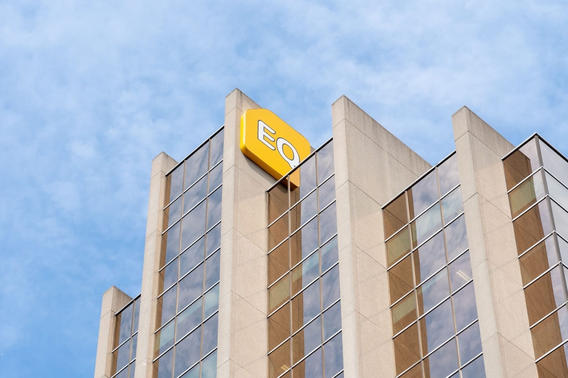 EQ Bank building with a yellow logo on the upper corner, featuring a modern glass and concrete facade against a partly cloudy sky.