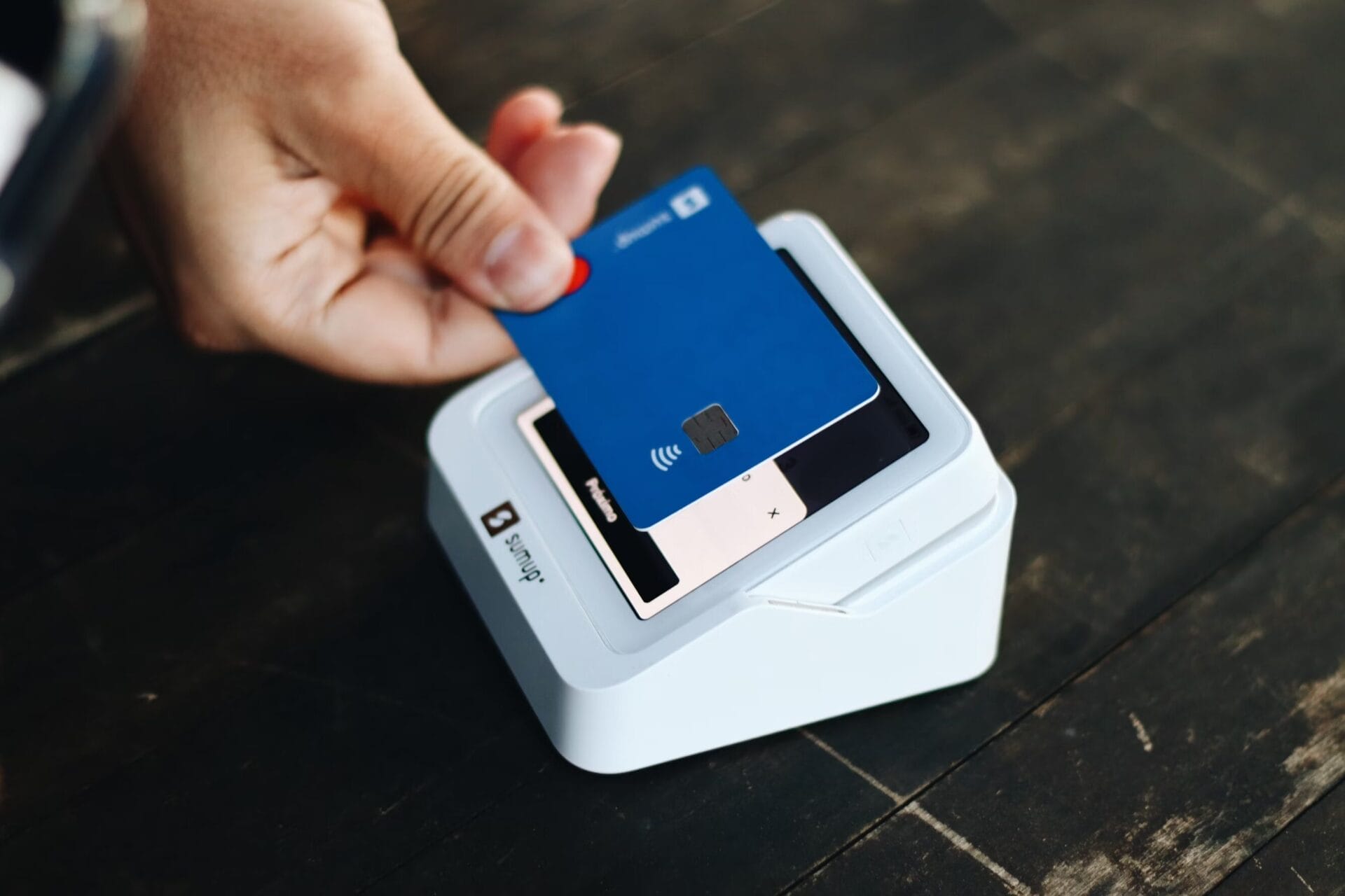 A close-up of a hand tapping a blue credit card on a white contactless payment terminal.