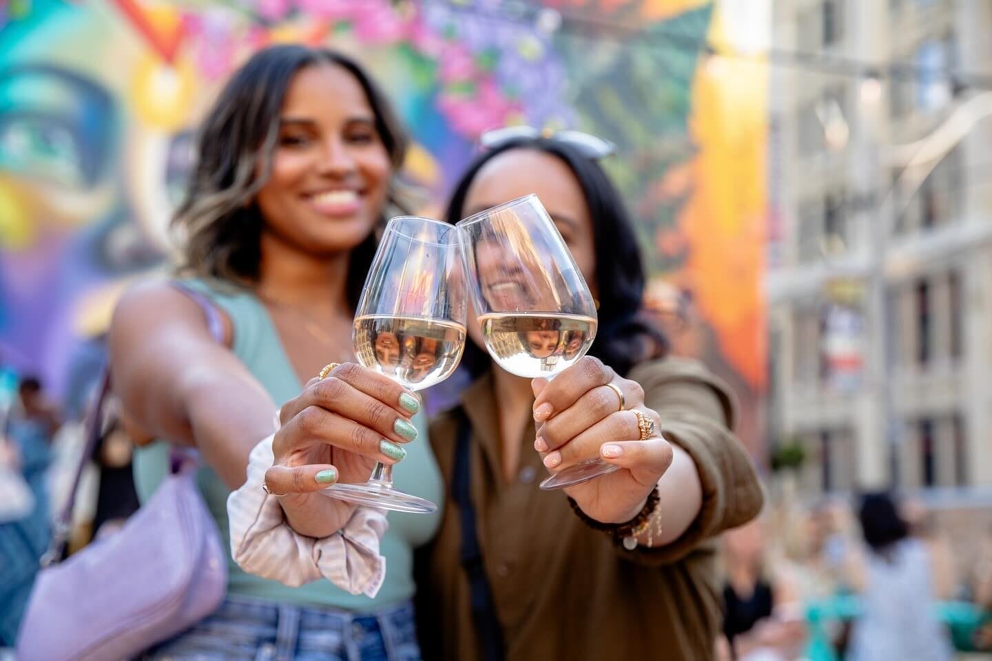 Two women clinking glasses of white wine, with a vibrant mural in the background.