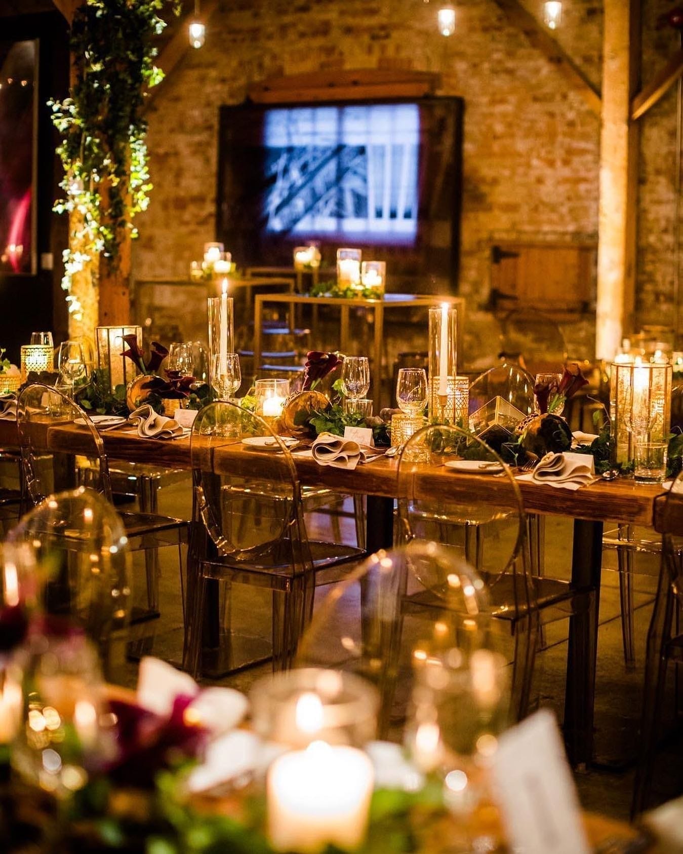 A beautifully decorated indoor dining area with rustic wooden tables, transparent acrylic chairs, soft candlelight, and elegant table settings. The warm, intimate ambiance features brick walls, hanging string lights, and lush green foliage used as table centerpieces and vertical garden accents.