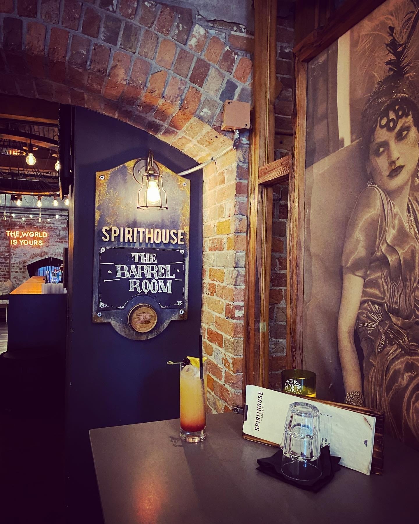 Cozy bar corner with a vintage vibe featuring ‘The Barrel Room’ sign, rustic exposed brick, warm lighting, a vintage portrait of a woman, and a refreshing cocktail.