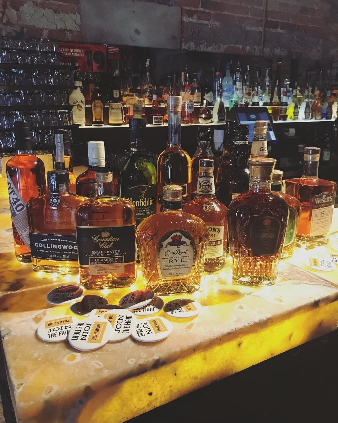 A selection of whiskey and spirits displayed on a backlit bar countertop, with a rustic brick wall and shelves of glassware in the background, evoking a warm and inviting atmosphere.