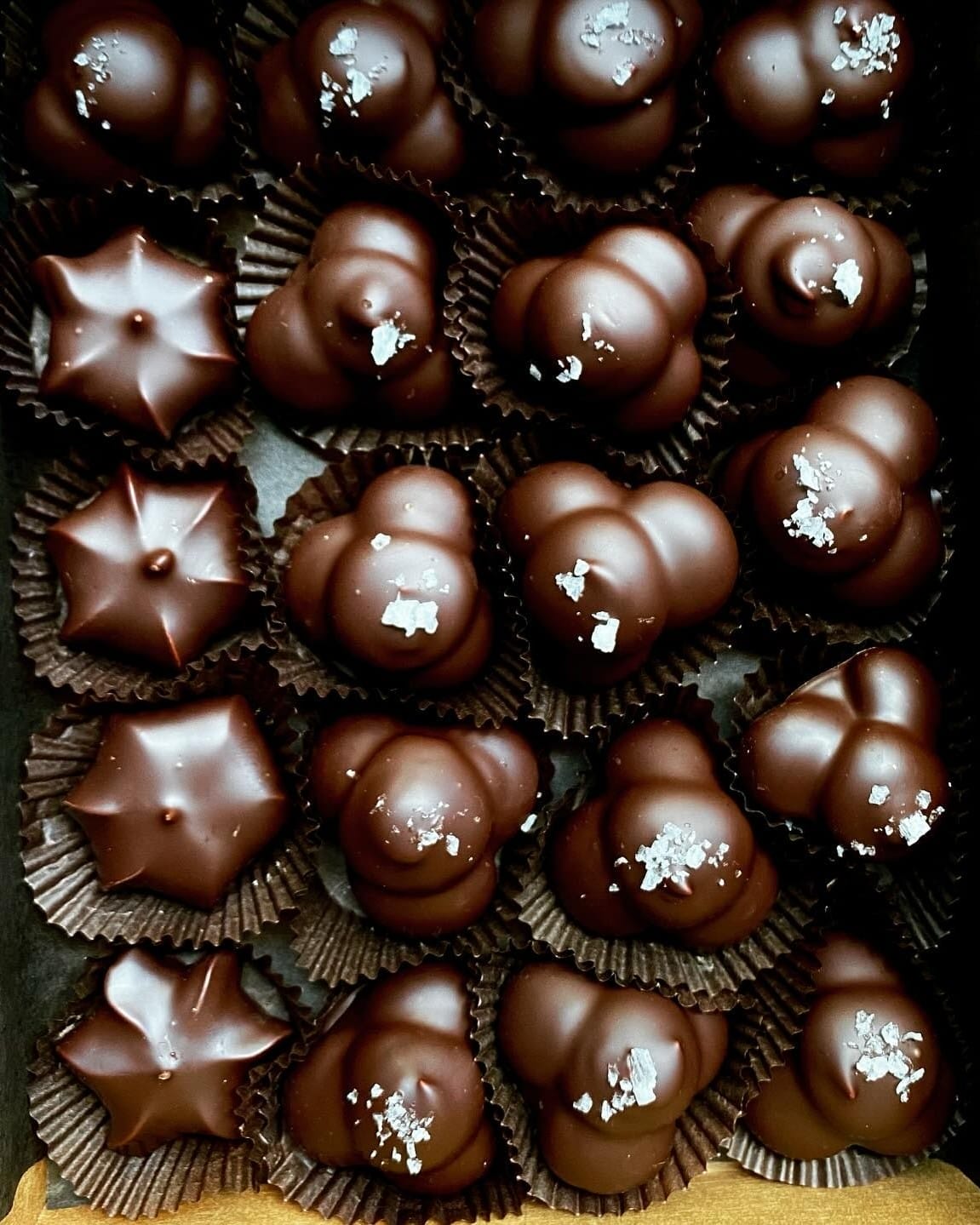 An assortment of glossy dark chocolate bonbons with unique shapes, some sprinkled with flaky sea salt, neatly arranged in a dark brown box lined with paper cups