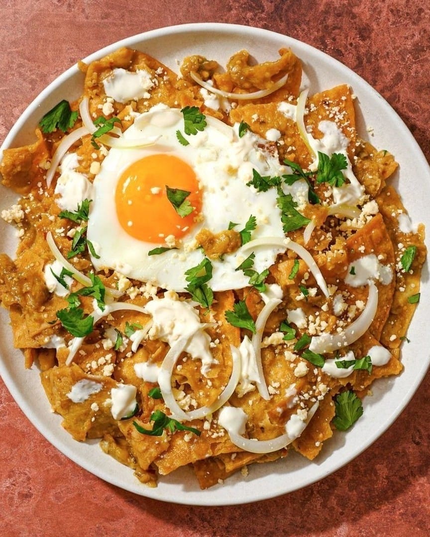 Plate of chilaquiles topped with a sunny-side-up egg, sliced onions, crumbled cheese, fresh cilantro, and a drizzle of crema.