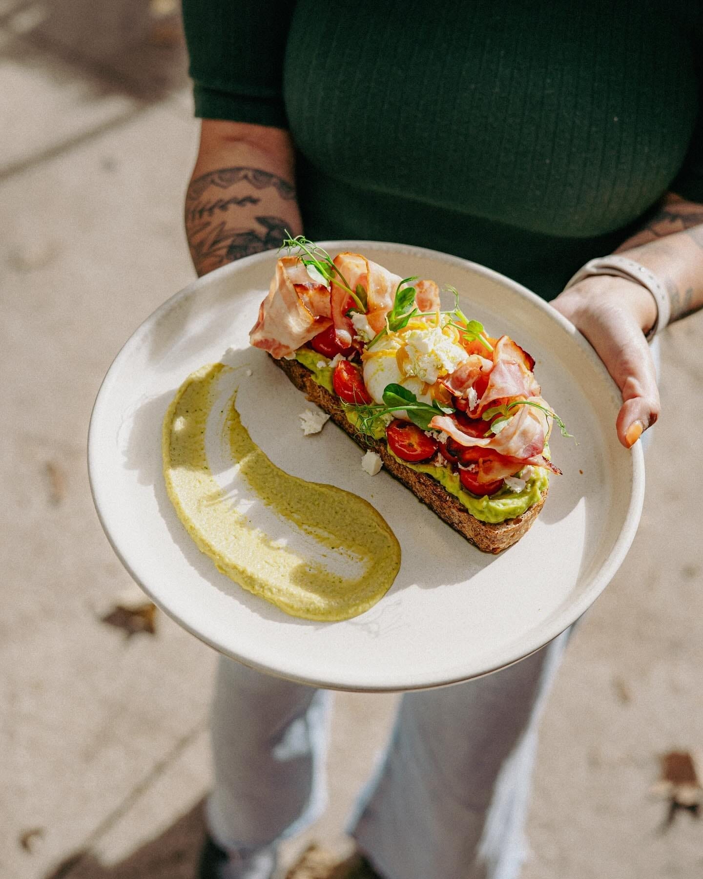 Hand holding a plate of gourmet avocado toast topped with poached egg, cherry tomatoes, crispy bacon, microgreens, and feta, served with a swirl of green sauce on the side.
