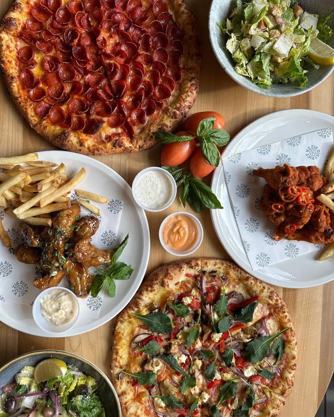 A table with pepperoni pizzas, salads, chicken wings, fries, and sauces on white plates, garnished with fresh herbs and tomatoes.