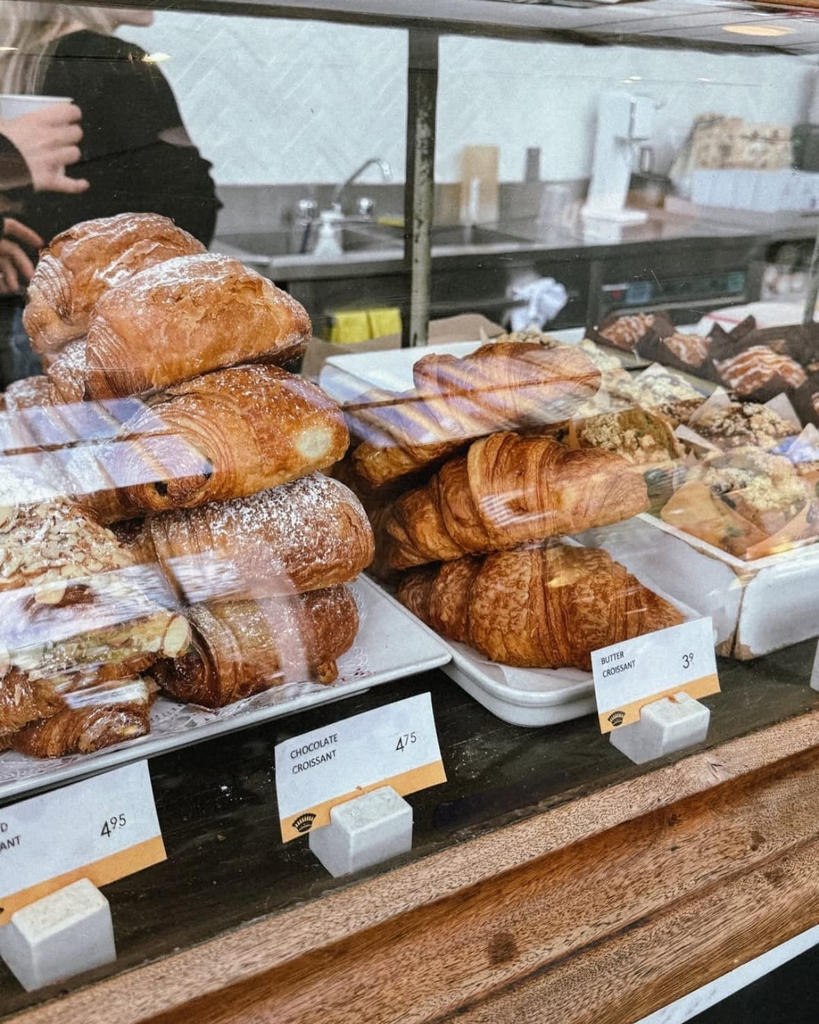 A close-up of golden, flaky croissants and almond pastries neatly stacked in a wooden display case at a bakery.