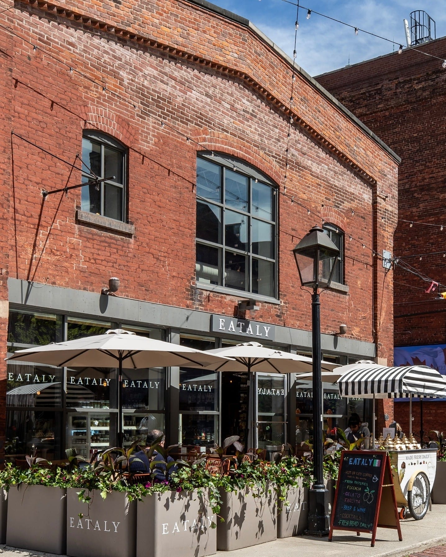 Outdoor patio of Eataly with white umbrellas and a black-and-white striped awning, featuring a chalkboard menu sign highlighting fresh offerings.