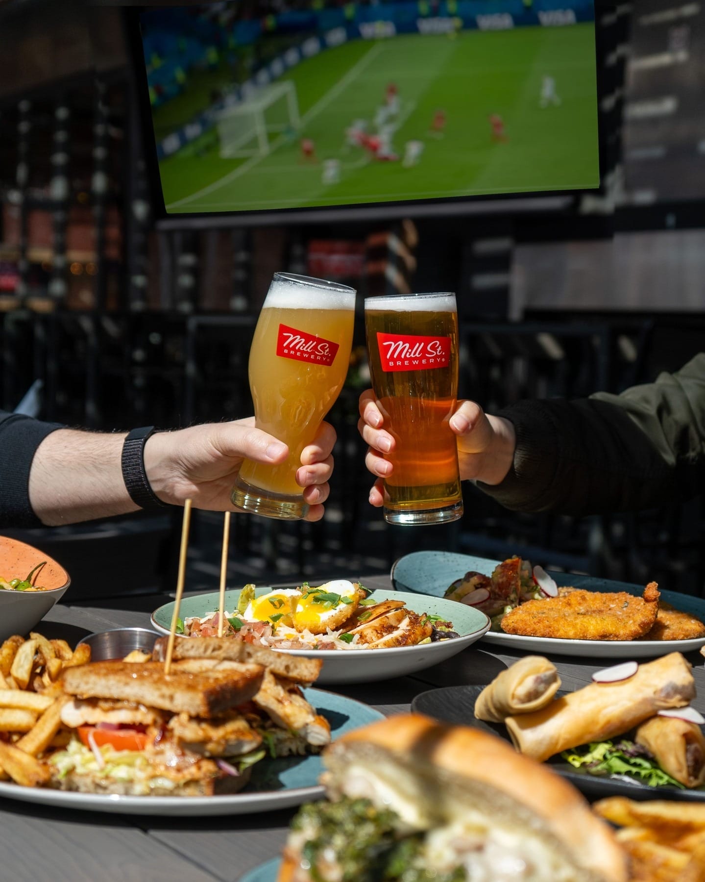 Friends enjoying pints of beer and hearty summer dishes on a sunny patio in Toronto’s Distillery Historic District, with a soccer match playing in the background