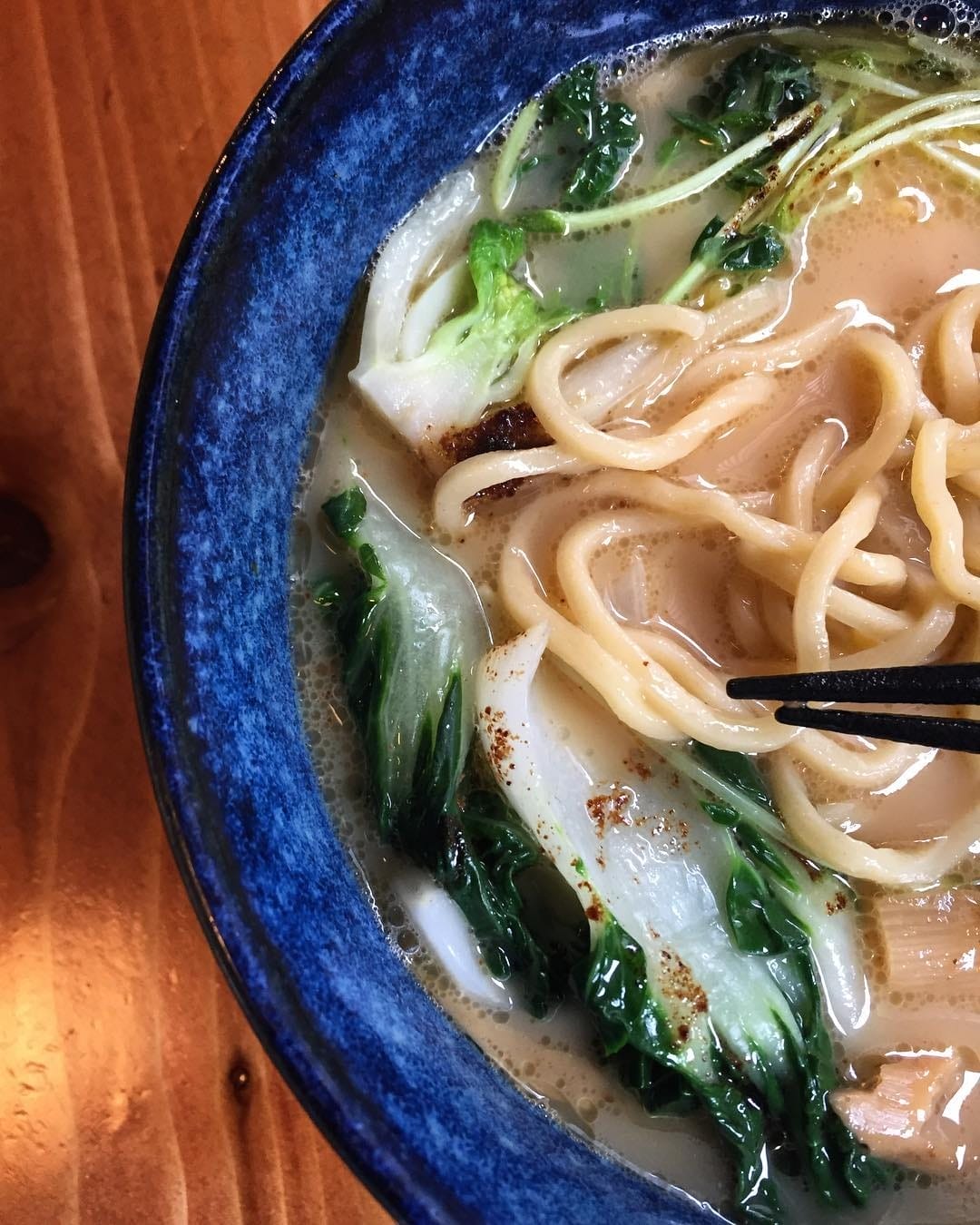 A steaming bowl of ramen with fresh greens and thick noodles, served in a deep blue ceramic bowl on a wooden table in Toronto’s Distillery Historic District