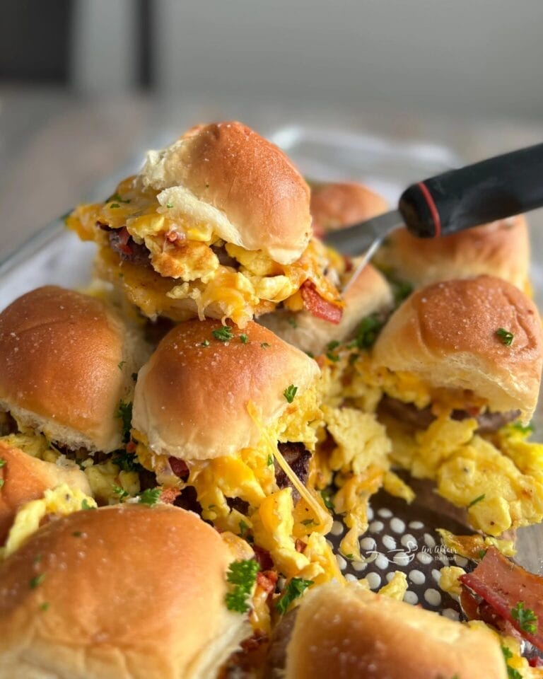 Close-up of cheesy breakfast sliders filled with scrambled eggs, sausage, and melted cheese, served on soft golden rolls in a glass baking dish.