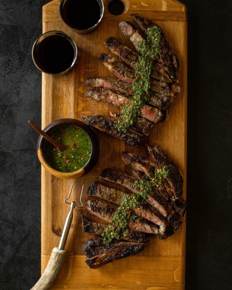Sliced grilled ribeye with chimichurri sauce served on a wooden board with red wine and herb sauce.