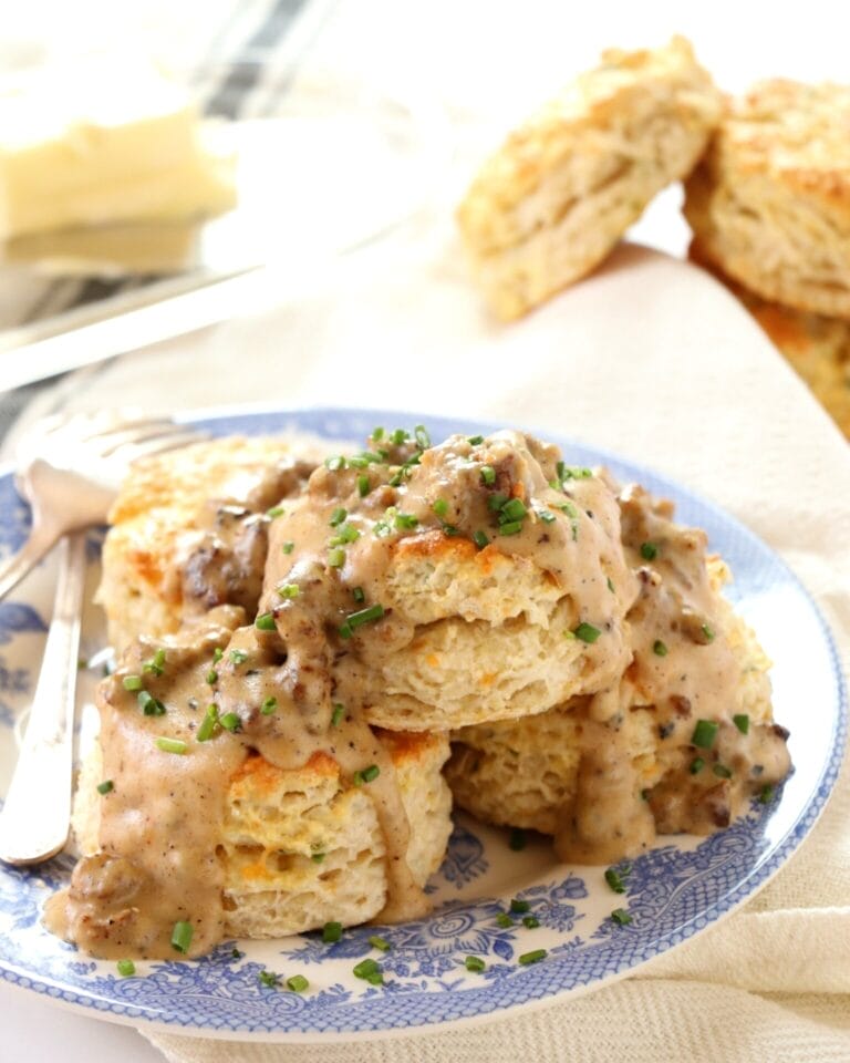 Flaky cheddar and chive buttermilk biscuits smothered in homemade sausage gravy, garnished with chopped fresh chives on a vintage blue-patterned plate.