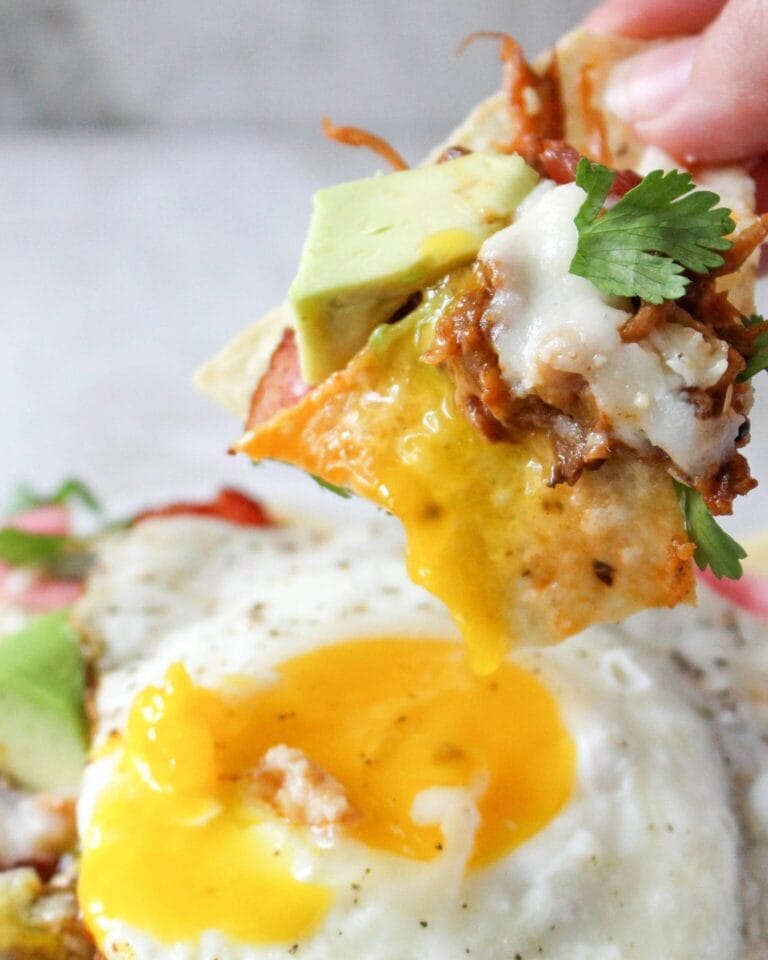 Close-up of a loaded breakfast nacho chip topped with melted cheese, pulled meat, avocado, cilantro, and a runny fried egg yolk in the background.