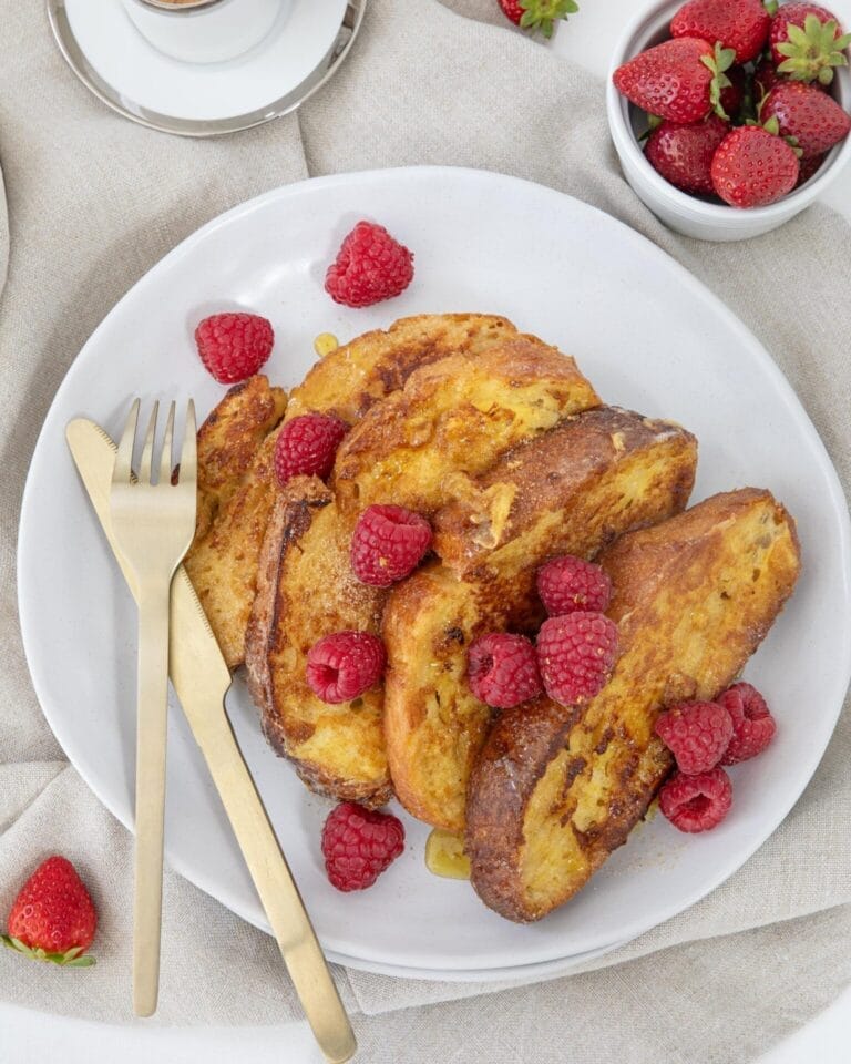 Golden brown sourdough French toast topped with fresh raspberries and a drizzle of syrup, served on a white plate with gold cutlery.